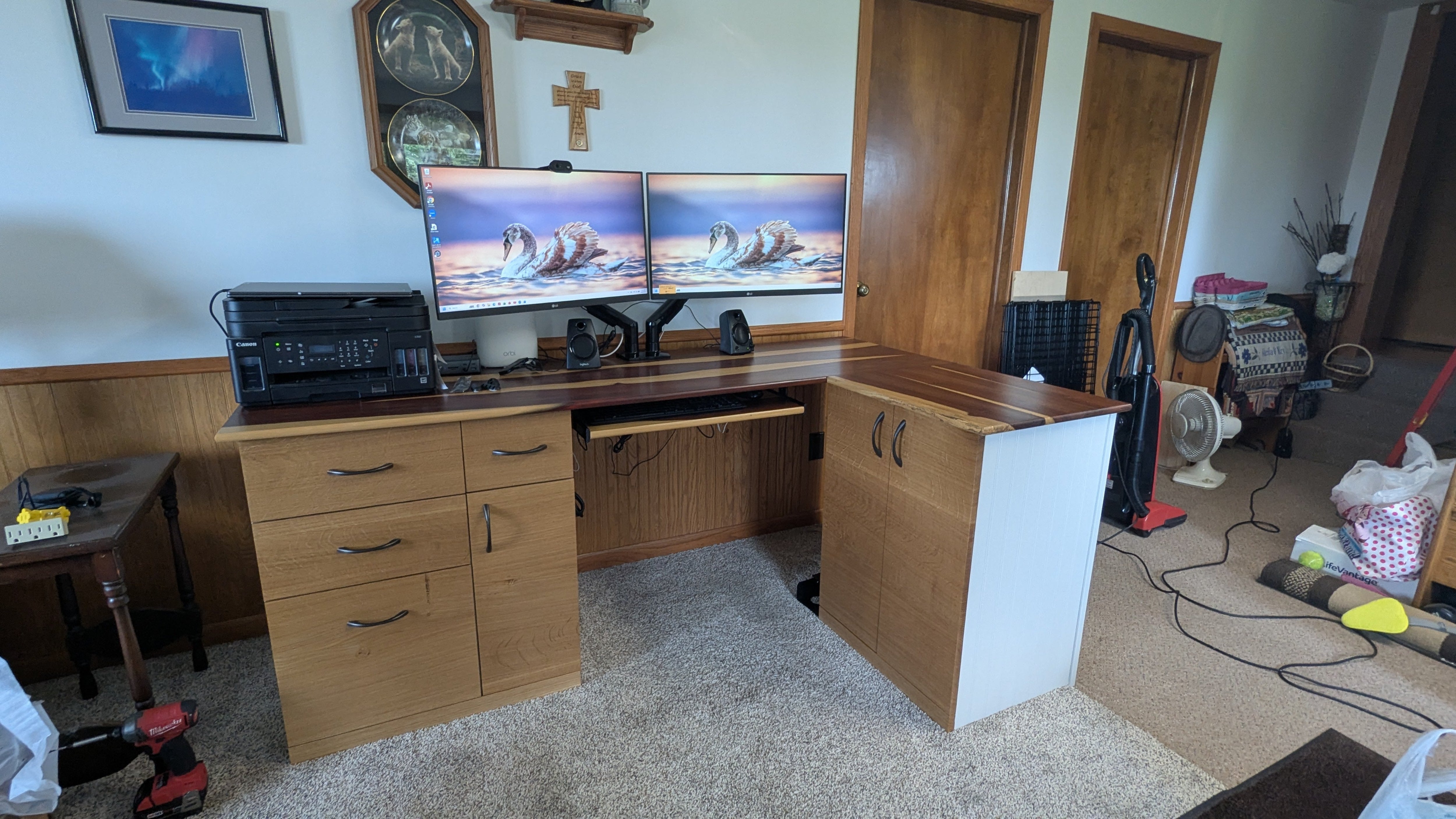 Wooden desk with computer setup in a room with carpeted floor and various items.